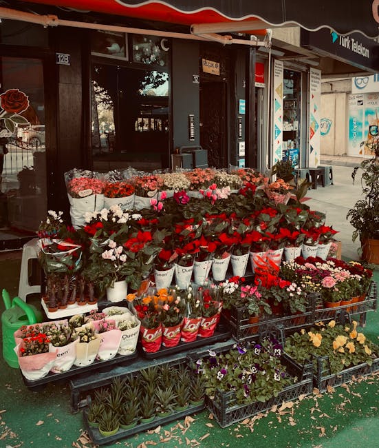 An outdoor display of fresh flowers in front of a flower shop, comprising various bouquets and potted plants arranged on black plastic crates and a table. The flowers include vibrant red roses, pink t