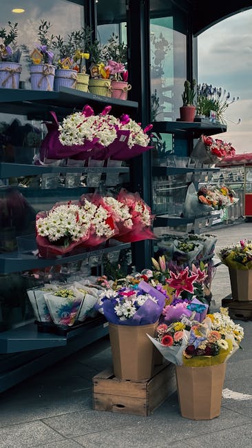 A display of fresh floral arrangements and bouquets arranged on dark grey metal shelves inside a flower shop with large windows, allowing natural light to illuminate the colorful flowers. The arrangem