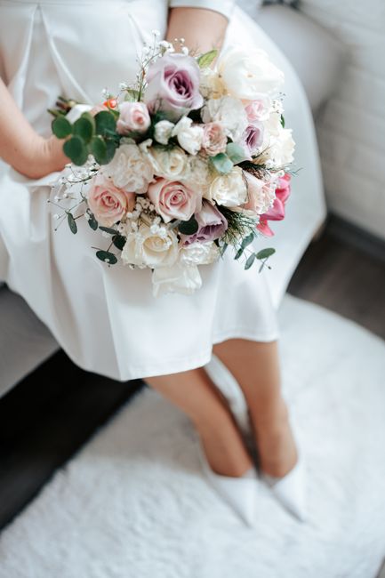 A person seated on a cream-colored sofa, holding a large, lush bridal bouquet with a mix of pastel pink and white roses, lavender-hued lisianthus, and sprigs of baby's breath, accented with green euca