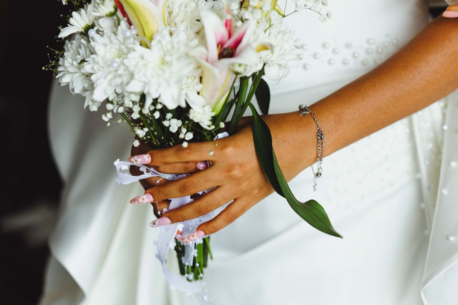 Close-up of a bride holding a bouquet of fresh white and pink flowers, including lilies, daisies, and baby's breath, wrapped in white ribbon with pearl accents. The bouquet is held in her left hand ag