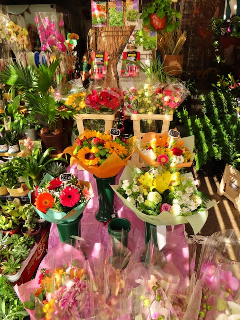 A vibrant display of fresh floral bouquets arranged on tall green stands and a pink table at a flower shop, with colorful flowers including pink, red, yellow, and white blooms such as gerbera daisies,