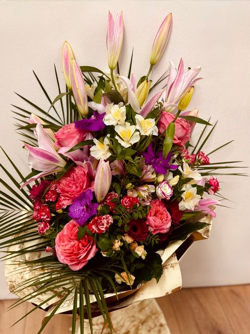 A vibrant floral arrangement featuring pink lilies with closed buds and elegant long stems, surrounded by pink roses, purple orchids, and small white and pink blossoms, all accented with lush green fo