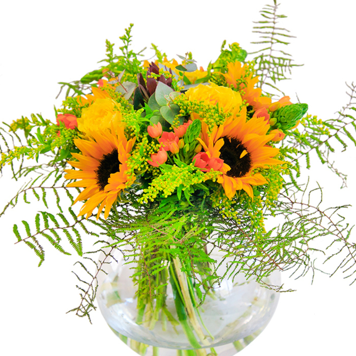Evoking the vibrant character of local Earlsfield gardens, this abundant bouquet is an uplifting testament to the skilled touch of community florists. Sunflowers, radiant with orange-yellow petals and bold dark centers, take pride of place-calling to mind golden rays splashed across the lawns of Garratt Green. Intermingled are classic yellow roses, their tightly wound petals adding a soft, luminous beauty and filling the air with a gentle fragrance. Scarlet Hypericum berries shine among the foliage, a playful accent that pops against the greenery and mirrors the lively spirit you might find at Kimber Road's weekend market. Feathery green ferns and delicate vines billow outwards, lending depth, movement, and the tranquil beauty of open spaces by the Wandle. Slim spikes of green and tiny yellow sprays suggest the last brilliance of summer meadows, while cool eucalyptus leaves introduce a calming, rustic touch. The round glass vase displays the crisscross of verdant stems in fresh water, a clean, modern finish that enhances the array's vivid palette. Whether shared at a festive dinner, gifted to mark a special moment, or simply enjoyed at home, this bouquet brings a radiant sense of optimism and Earlsfield soul to every occasion, beautifully embodying the story of local blooms.